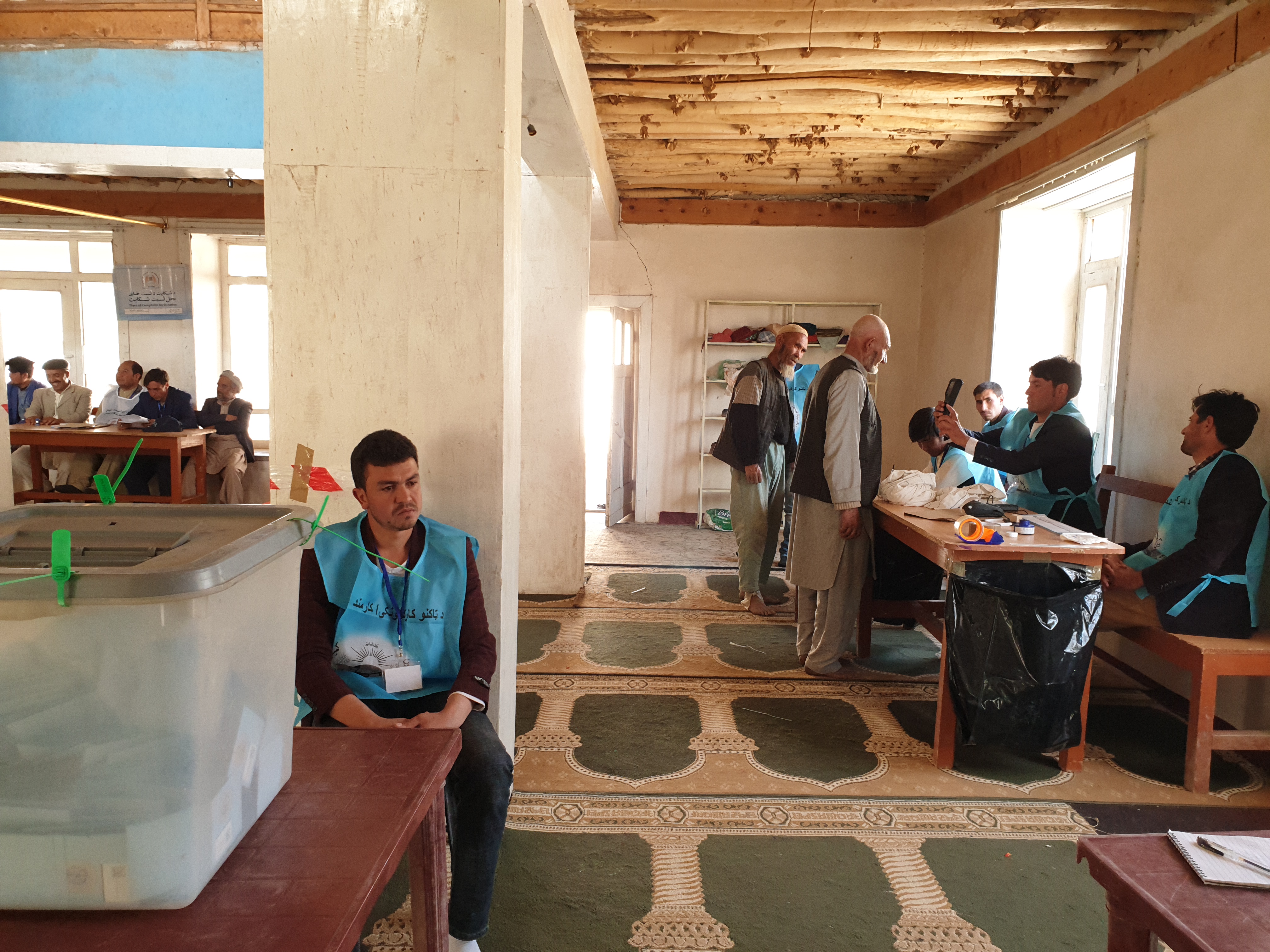 A BVV operator takes a photo of a voter’s face, while a ballot controller looks on, the ballot box controller waits and a group of agents and observers look on at the Shinya Madrasa in Foladi, Bamyan. Photo: Ali Yawar Adili, 28 September 2019